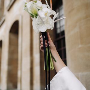 Modern white bouquet featuring anthuriums, orchids, and calla lilies with black ribbon, held up against a stone building backdrop.