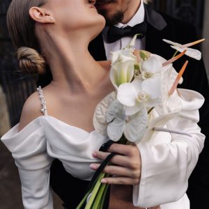 Bride and groom embracing with a modern white bouquet of orchids, anthuriums, and calla lilies tied with black ribbon.