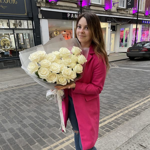 Woman in a bright pink coat holding a bouquet of white roses from KS Bloomery, standing on a stylish UK high street.