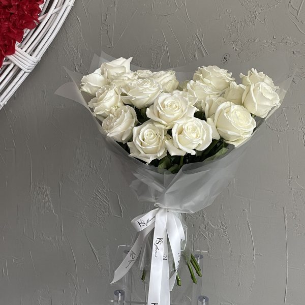 Elegant bouquet of white roses wrapped in translucent paper and tied with a white KS Bloomery ribbon, set against a grey textured wall.