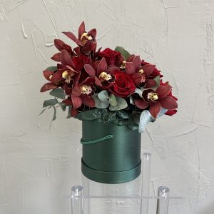 Luxury floral arrangement with deep red roses and burgundy orchids in a green hat box.