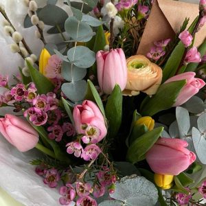 Close-up of a spring bouquet with pink tulips, yellow ranunculus, waxflowers, eucalyptus, and pussy willow branches, with a kraft paper envelope tucked in.